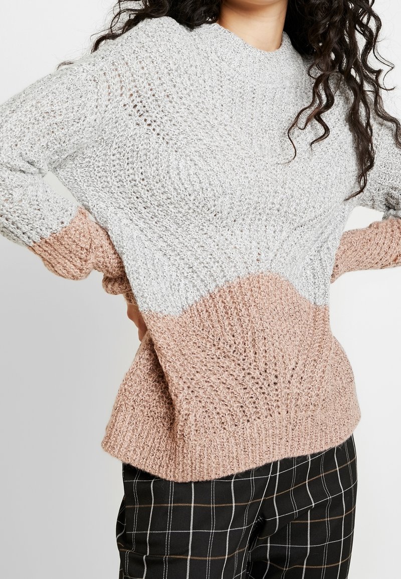 Femme portant un pull en maille texturée avec des sections supérieures grises et inférieures marron clair, associée à un pantalon à carreaux noir, mains sur les hanches.