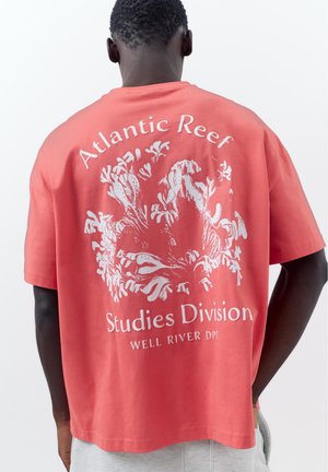 Homme portant un t-shirt corail avec un motif récif corallien blanc et le texte « Atlantic Reef Studies Division Well River Dpt » dans le dos.