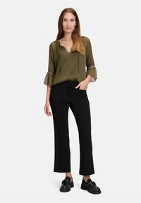Blusa verde oliva con scollo a V e cordoncino, abbinata a pantaloni neri a flare e scarpe platform nere chunky. Maniche semi-trasparenti.
