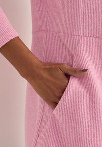 Pink ribbed fabric dress with long sleeves, featuring a side pocket. Close-up of a hand resting on the pocket. Soft texture evident.