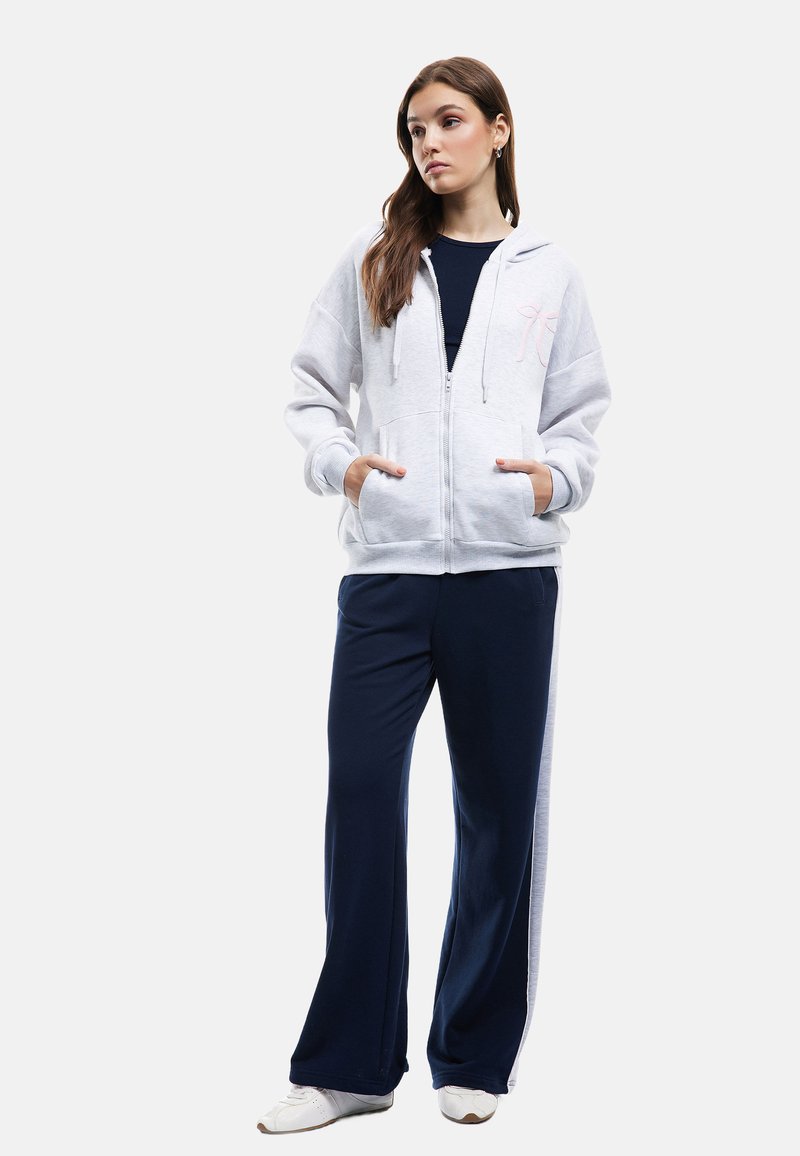 Lys grå zip-up hættejakke med pink broderi, kombineret med mørkeblå brede bukser og hvide sneakers. Glat stof med lommer.
