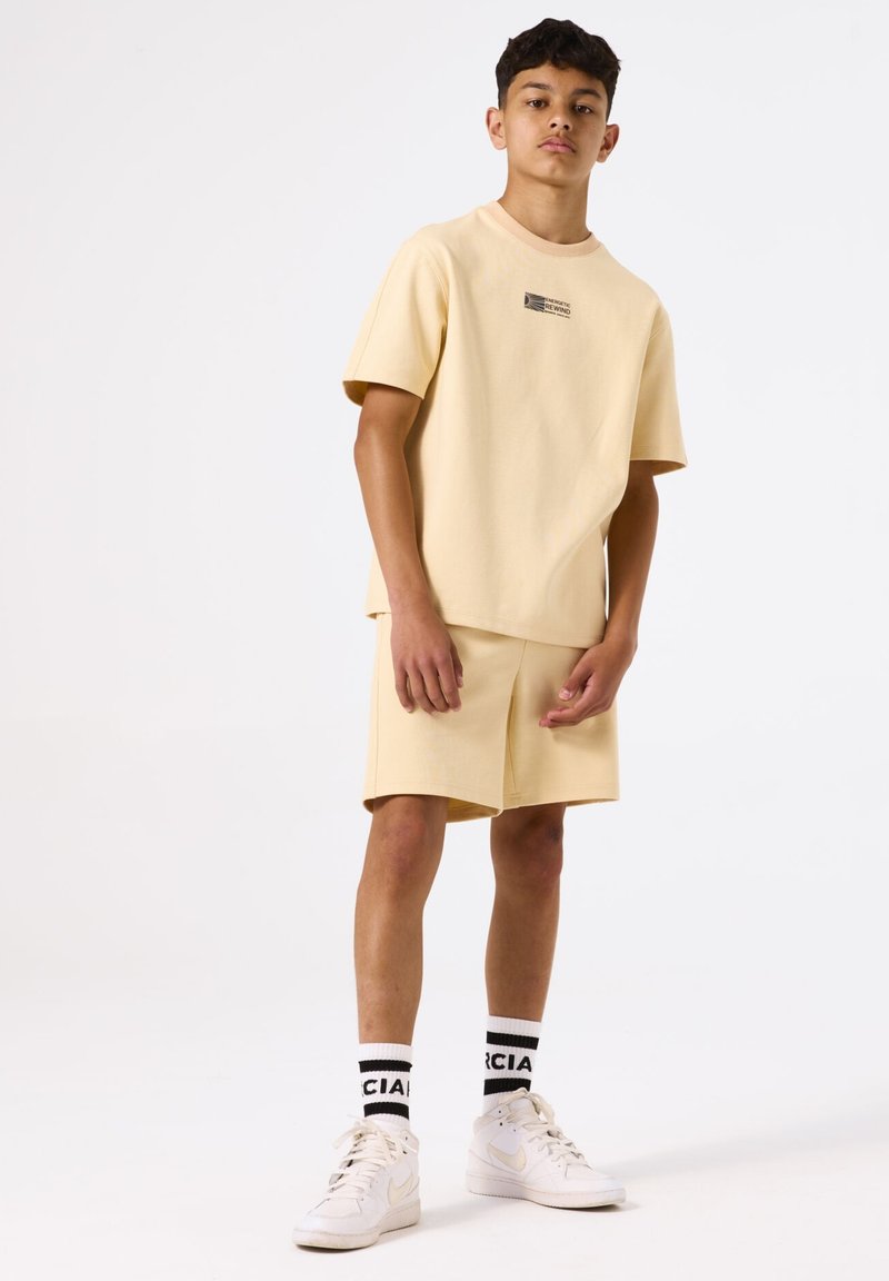 Geel katoenen t-shirt met een klein logo, bijpassende shorts, witte sneakers en zwart-witte merk sokken met horizontale strepen.