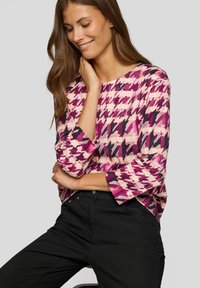 Frau mit langem braunem Haar, die lächelt, trägt ein pink-schwarz gemustertes langärmliges Oberteil und schwarze Hose vor einem hellgrauen Hintergrund.