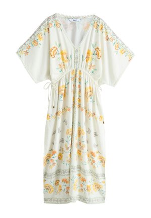 Kvinde med krøllet hår iført en hvid blomsterprintet maxikjole med brede ærmer, stående på stentrapper ved en hvid væg.