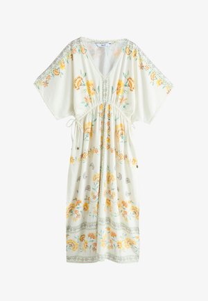 Frau mit lockigem Haar, die ein weißes, florales Maxikleid mit weiten Ärmeln trägt, steht auf Steinstufen vor einer weißen Wand.