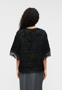 Μαύρο τοπ με κρόσσια και στρογγυλό ντεκολτέ, διακοσμημένο με υφή σε όλη την επιφάνεια και κοντά μανίκια, φορεμένο πάνω από γκρι ένδυμα.