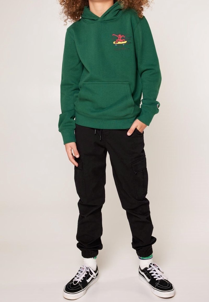 Zelená mikina s kapucí a přední kapsou, se skateboardovým grafickým potiskem ve stylu graffiti, v kombinaci s černými cargo kalhotami a černými teniskami s bílými akcenty.