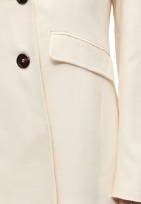 Blazer en tissu texturé crème avec des boutons brun foncé et une poche à rabat visible sur le côté droit près de la manche.