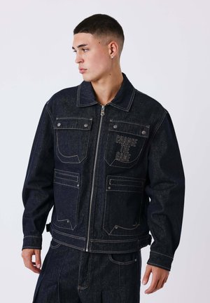 Jeune homme portant une veste en jean foncé et un pantalon, la veste a des poches avant et le texte "Change the Game" sur la poitrine gauche, regardant vers sa gauche.