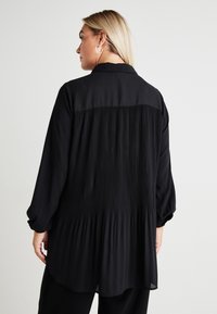 Black blouse featuring a pleated back design, long sleeves, a collared neckline, and a slightly elongated hem for a relaxed fit.