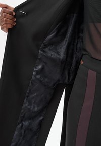 Hand die een open zwart jack vasthoudt om de binnenvoering met een donker paisley-patroon te tonen, naast een persoon die zwarte broek draagt met een donkerpaarse streep.