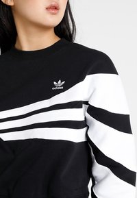 Black sweatshirt with white stripes and an embroidered Adidas logo on the chest. Features a crew neckline and a soft, textured fabric.