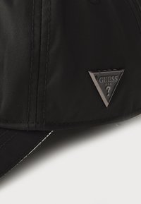 Căciulă de baseball neagră din nylon cu o textură netedă, având un badge triunghiular negru embosat cu „GUESS USA”. Accente de cusături albe.
