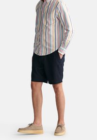 Chemise à rayures à manches longues avec des couleurs pastel, shorts navy et chaussures beiges. La chemise possède une poche avant et des manches retroussées.