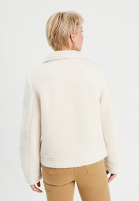 Veste en shearling créme, moelleuse, avec un large col, des épaules drop et une coupe courte. Tissu texturé et détails cousus visibles.