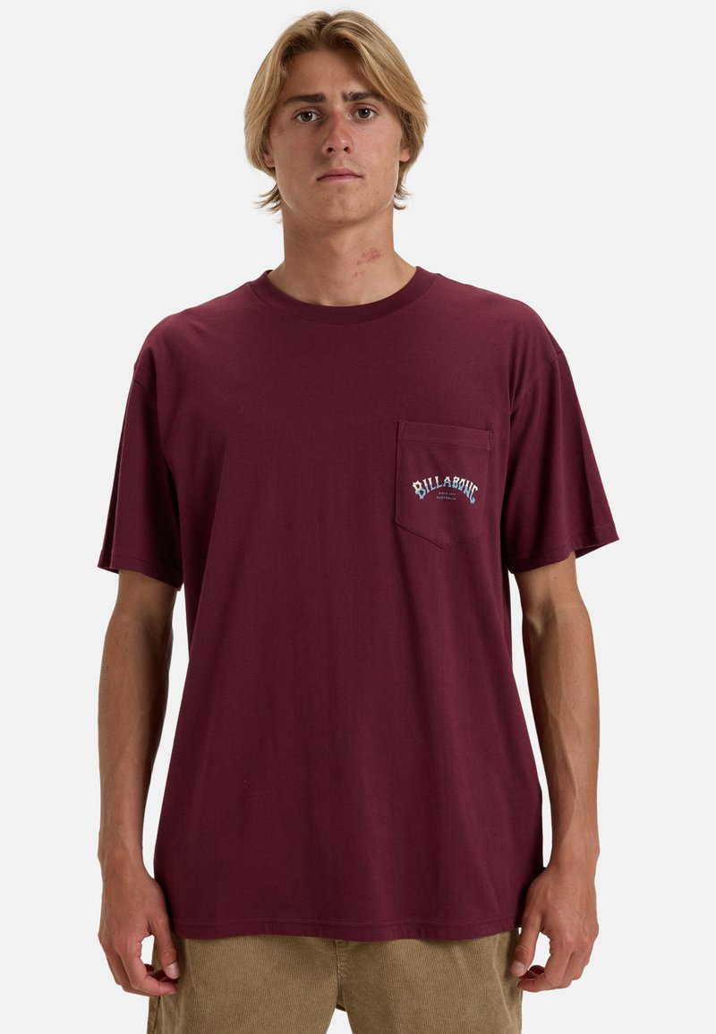 Bordeauxfarbenes T-Shirt mit kurzen Ärmeln aus Baumwolle, ausgestattet mit einer Brusttasche und dem weißen "Billabong"-Logo. Lockerer Schnitt, glatte Textur.
