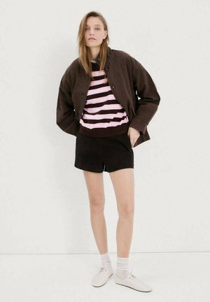 Giovane donna con giacca marrone, maglione a righe rosa e marrone, pantaloncini neri, calzini bianchi e scarpe bianche, in piedi contro un muro bianco.