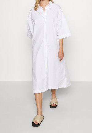 Frau geht und trägt ein locker sitzendes weißes Midi-Kleid mit Knopfleiste, Ellbogenlangen Ärmeln und beigen Schlupfsandalen.