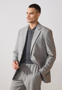 Grauer maßgeschneiderter Blazer über einem dunkelgrauen Poloshirt, kombiniert mit grauen Hosen mit elastischem Bund. Glattes Material, strukturiertes Design und subtile Details.