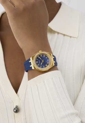 Guldtonet ur med blå urskive og romerske tal, pyntet med krystaller. Har et blåt silikonerem og et ottekantet kassedesign.