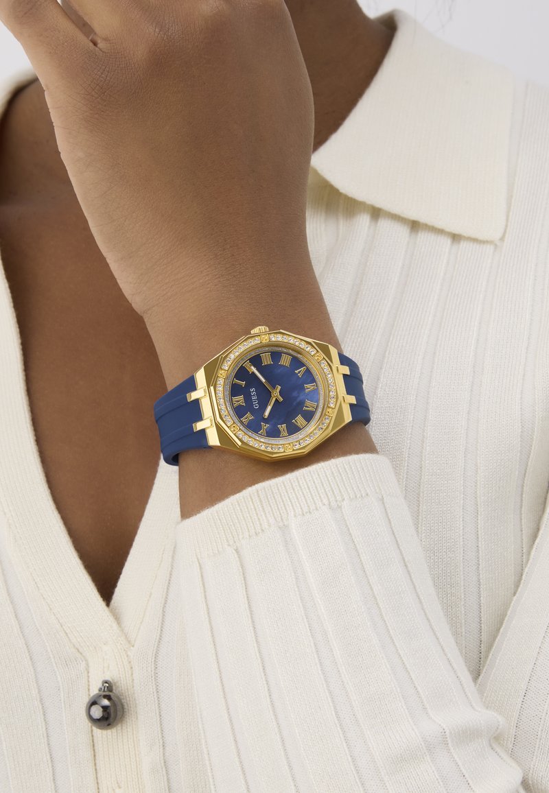 Złoty zegarek z niebieską tarczą i cyframi rzymskimi, ozdobiony kryształami. Posiada niebieski pasek silikonowy i ośmiokątną obudowę.