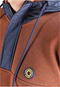 Gros plan d'une veste marron texturée avec un col matelassé bleu marine, un cordon de serrage et un patch rond avec le logo "GABBIANO" près de la poche poitrine.