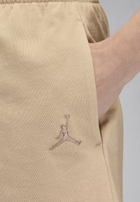 Kreminės spalvos sportinės kelnės, pagamintos iš minkštos medžiagos, su matoma kišene, šone turi smėlio spalvos siuvinėtą Jumpman logotipą.