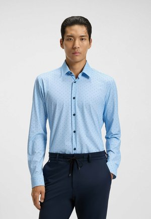 Chemise bleu clair à boutons avec de petits pois noirs, manches longues, un col et des boutons noirs. Portée avec un pantalon sombre.