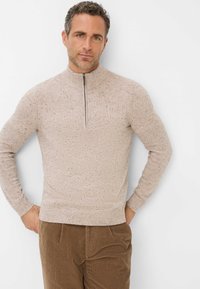 Helles beige Pullover mit hohem Kragen, Frontreißverschluss und gesprenkelter Textur, kombiniert mit braunen Cordhosen.