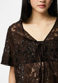 Bruin kanten topje met bloemmotieven, pailletten en een striksluiting. Het ontwerp heeft korte mouwen en een diepe V-hals.
