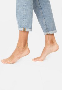 Kuldne sääreketti paljal jalal, keritud helesiniste denim teksadega. Ketil on väikesed ümmargused lingid. Pehme tekstuur on selgelt nähtav.