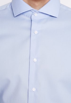 Camicia azzurra chiaro con bottoni, realizzata in tessuto liscia. Presenta un colletto classico e sei bottoni sul davanti. Tessuto uniforme, senza pattern visibili.