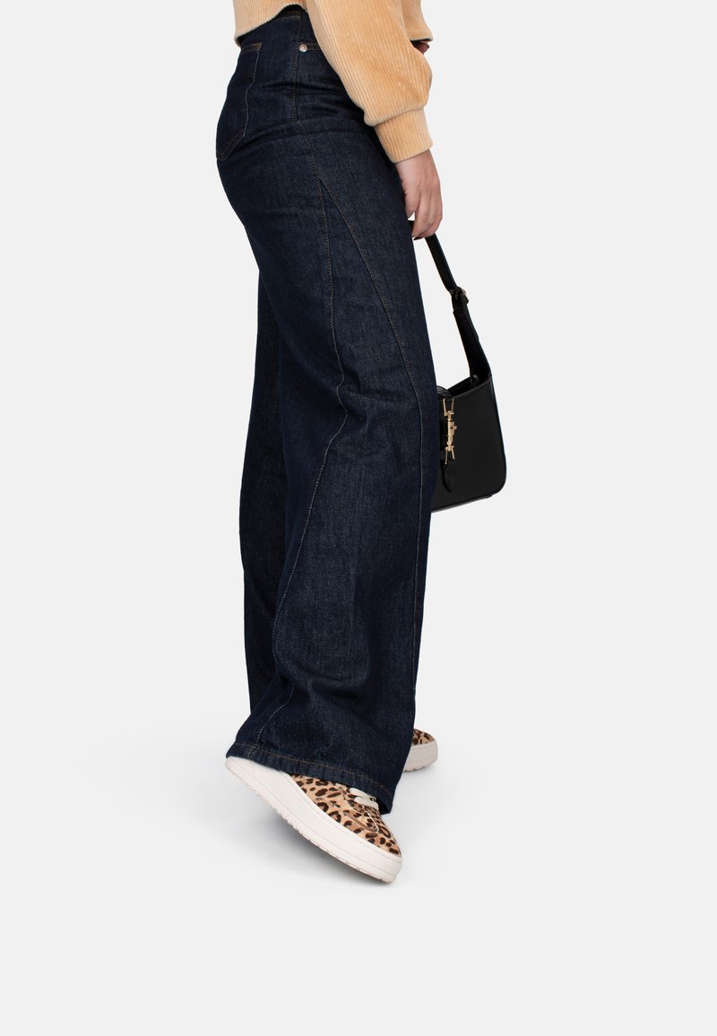 Weite dunkelblaue Denim-Jeans mit sichtbaren Nähten, kombiniert mit Sneakers im Tiermuster und einer kleinen schwarzen Handtasche.