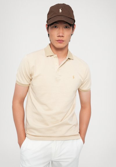 Beige polo skjorte med et teksturert mønster og en kontrasterende krage, kombinert med en brun caps med et logo. Modellen står med hendene i lommene.