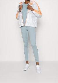 Veste coupe-vent blanche avec des accents orange, haut ajusté bleu clair, et leggings bleu clair à côtes. Des baskets blanches complètent cette tenue sportive.