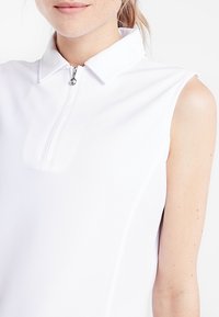 Bílý bezrukávový polo s stojáčkem, má přední zip a strukturovanou tkaninu. Minimalistický design, vhodný pro aktivní nošení.