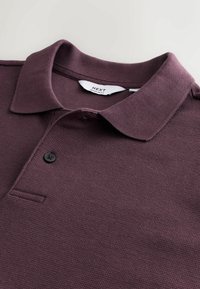Lähellä kuva marooninvärisestä texturoituneesta polo-paidasta, jossa on laskettavat kaulukset, yksi näkyvissä oleva musta nappi ja "NEXT Regular Fit" -labeli kauluksessa.