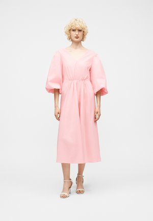 Personne aux cheveux blonds bouclés portant une robe midi rose à manches bouffantes et des sandales blanches à talons hauts, sur un fond clair uni.