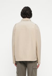 Lühipäine inimene, seljas beež North Face'i jakk ja oliivrohelised cargo-püksid, on nähtav seljaga lihtsa valge tausta ees.