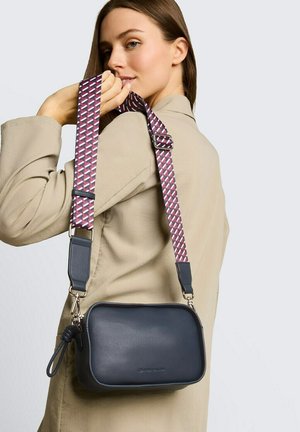 Femme en manteau beige tenant un sac bandoulière bleu marine avec une large sangle à motif sur l'épaule, devant un fond clair.