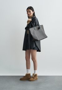 Black and brown tote bag with a geometric pattern, featuring two black handles. Model wears a black button shirt and brown ankle boots.