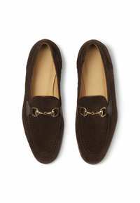 Mocasines de pana marrón con una correa de cuero suave y detalle en metal dorado en la parte superior. Diseño de punta redondeada. Forro interior suave.