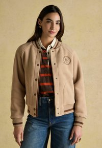 Beež varsity jakk, millel on pruunid triibud, nööpideks nupud ja broderitud logo. Kandmiseks triibulise särgi peal ja denimitest.