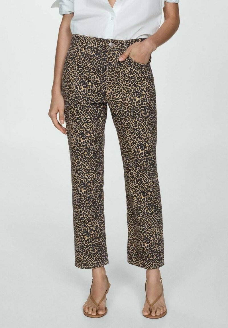 Femme portant un pantalon court imprimé léopard, une chemise blanche boutonnée et des sandales marron à brides, debout avec une main dans la poche.