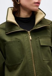 Γυναίκα που φοράει μπουφάν ελιάς με γιακά επενδεδυμένο με μπεζ, χρυσό φερμουάρ, τσέπες με καπάκι και επιθέματα, και χρυσό σκουλαρίκι σε σχήμα σταγόνας, εμφανιζόμενη από το λαιμό έως το στήθος.