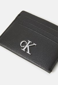 Tarjetero de cuero negro con tres ranuras para tarjetas y un logo plateado "CK". La superficie tiene un acabado texturizado. Forma rectangular compacta.