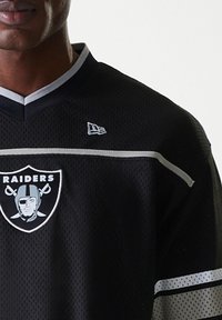 Maillot de football noir avec tissu en maille, portant un logo des Raiders, des accents argentés et un design en V. Le haut présente une bande horizontale gris clair.