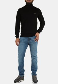 Maglione dolcevita a righe nere abbinato a jeans in denim azzurro chiaro, completato da sneakers scure, con un accento discreto del logo verde.