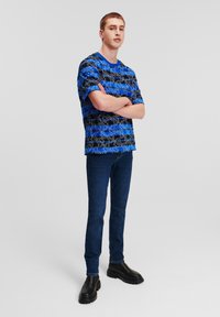 Rövid ujjú kék ing fekete és fehér absztrakt mintákkal, sötétkék, slim-fit farmernadrággal és fekete, vastag talpú csizmával párosítva.