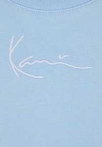 Tissu en coton bleu clair avec un motif de signature brodé en blanc. Texture lisse, encolure arrondie, et sans motifs ni accents supplémentaires.